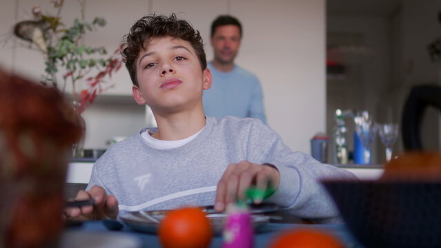 Boy sitting at dining table during family meal, looking ahead with content expression while food and fruit are placed on table in cozy home kitchen