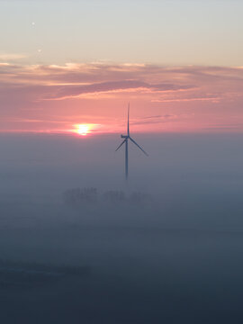 Aerial view of a single wind turbine rising above a thick layer of morning fog under a vibrant pink and orange sunrise sky in Vrouwenpolder, Zeeland, Netherlands.