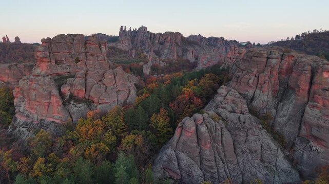 Medium-wide aerial pass along natural stone cliffs above dense autumn woodland in soft evening light.