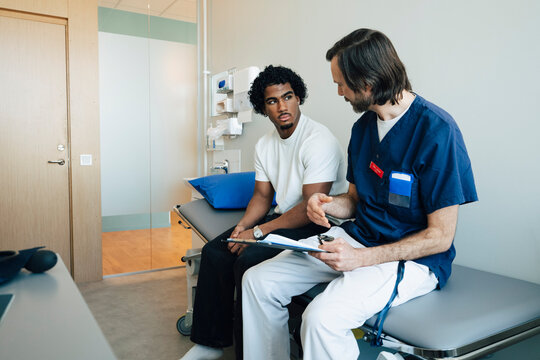Young man listening to male doctor explaining medical records while sitting on examination table in room at hospital