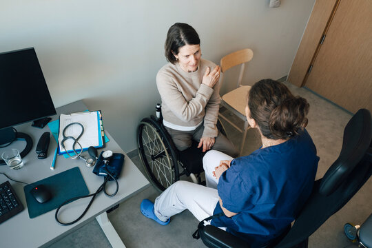 High angle view of woman sitting on wheelchair explaining shoulder pain to female physician during visit at hospital