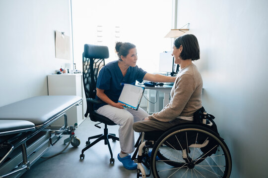 Happy female healthcare professional sharing positive medical records to woman sitting on wheelchair in examination room