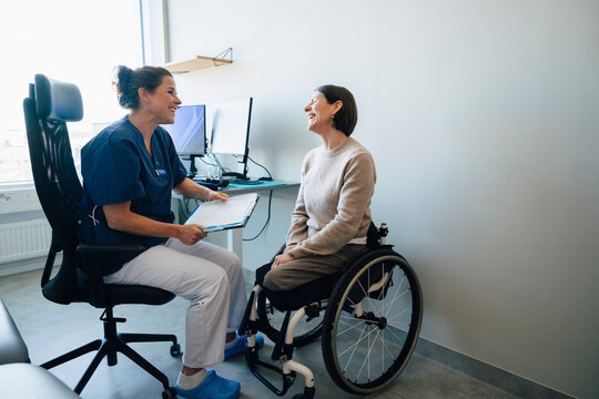 Happy woman sitting on wheelchair talking with female doctor sharing medical record with her in hospital room