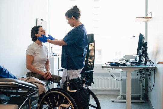 Female medical expert checking throat of mature woman having physical disability sitting on examination table in clinic