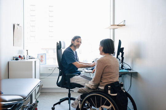 Male doctor listening to mature woman sitting on wheelchair during visit in hospital room