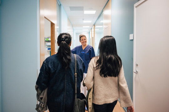 Rear view of mother and daughter walking towards female doctor standing outside examination room in hospital