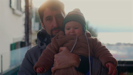Father making playful face while holding baby in bear teddy fleece coat and striped beanie hat outdoors, man bonding with child during joyful cold season family outing © Marco
