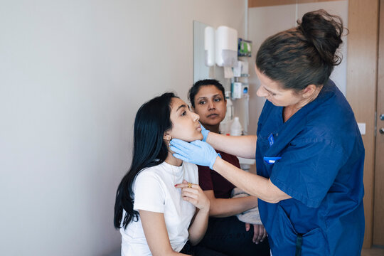 Female medical professional examining throat of girl sitting in examination room with mother at hospital