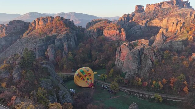 Wide aerial view of a hot air balloon beside dramatic rock formations, autumn trees, and an open launch site.