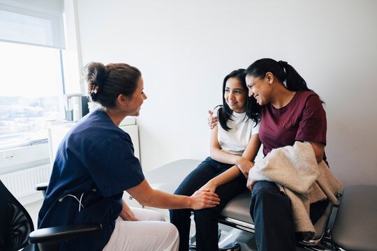 Smiling female pediatrician consoling girl getting embraced by mother in hospital room