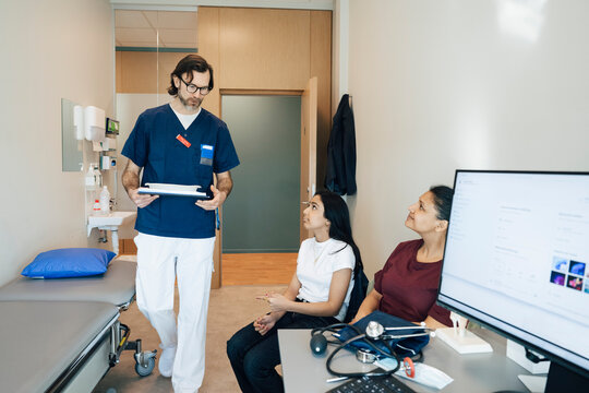 Male pediatrician walking near mother and daughter sitting in room during hospital visit
