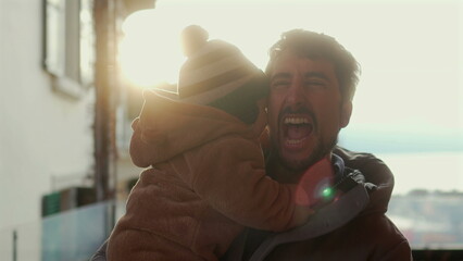 Father holding baby in bear teddy fleece coat and striped green beanie hat outdoors, man smiling with joy as infant hugs his face during warm and affectionate winter moment © Marco