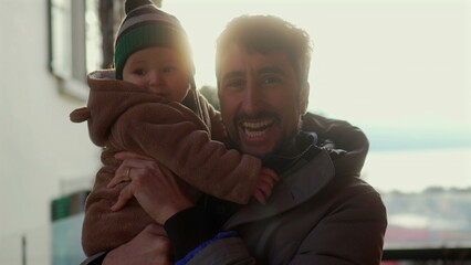 Father holding baby in bear teddy fleece coat and striped green beanie hat outdoors, man smiling with joy as infant hugs his face during warm and affectionate winter moment © Marco