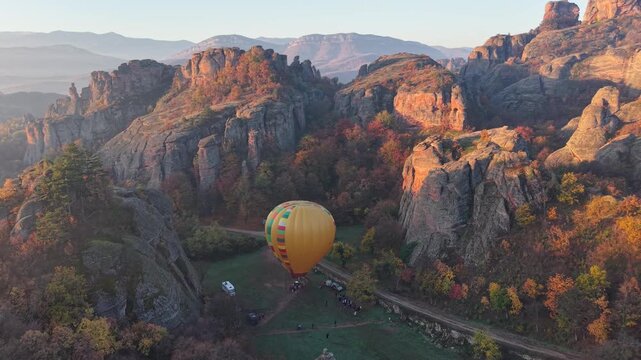 Wide aerial view of a hot air balloon on an open launch field surrounded by rocky cliffs and autumn trees.