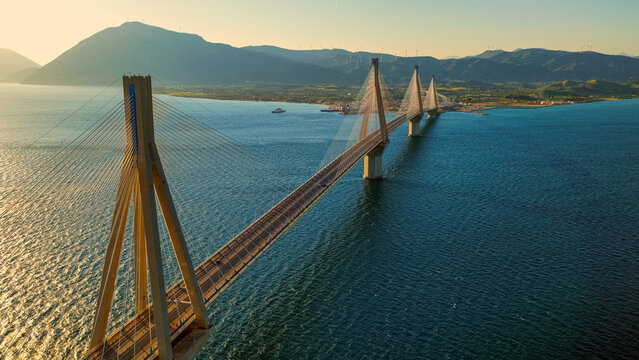 Rio&ndash;antirrio bridge spans the blue gulf of corinth at golden hour, cable-stayed landmark connecting peloponnese to mainland greece with distant mountains and wind turbines