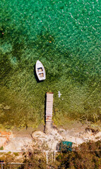 Fototapeta premium Small white rowboat floating on clear turquoise water near a rustic wooden pier and rocky shoreline, providing a peaceful scene for summer vacation or holiday