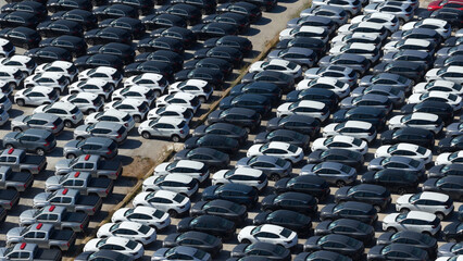 Rows of new silver, white, and black cars, including sedans, suvs, and pickup trucks, parked tightly on a spacious outdoor market lot, showing automotive industry logistics and inventory management © Nasos Zovoilis