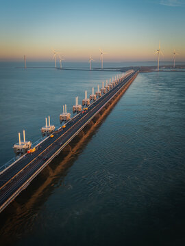 Aerial view of the Oosterscheldekering storm surge barrier with wind turbines and a coastal road during sunset in Vrouwenpolder, Zeeland, Netherlands.