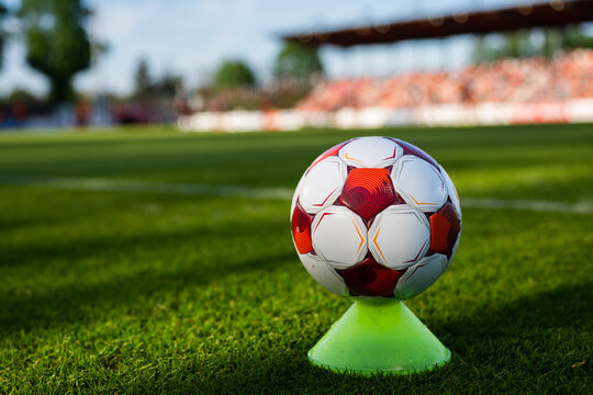 Red and white soccer ball placed on a  training cone on a grass pitch with stadium seating in the background.