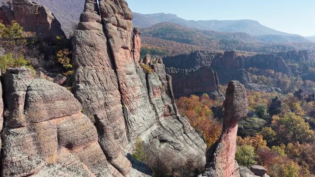 Close aerial pass along natural stone cliffs and a tall rock pillar above colorful woodland.