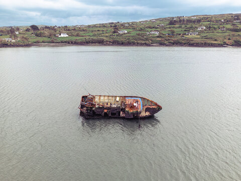 Aerial view of a rusty shipwreck abandoned in shallow coastal waters with green hills and houses in the background under an overcast sky.