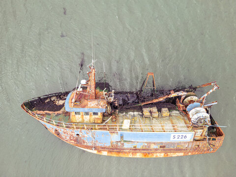 Aerial view of a rusty abandoned fishing boat with registration S 226 resting in shallow murky water with seaweed visible on the deck.