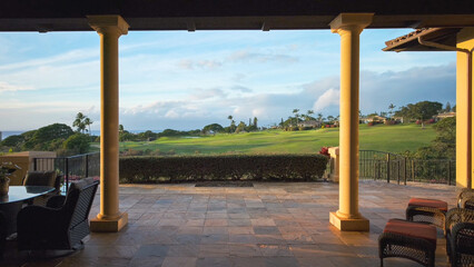Fototapeta premium View from the terrace of a luxury house with large pillars that offers a view of the expansive green golf course and the sea in the distance at sunset