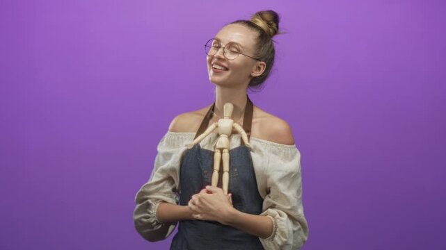 Woman in apron hugging a wooden mannequin to bare shoulders, smiling and looking up in a purple studio; creative joy.
