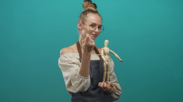 Woman blonde wearing glasses and denim apron holds a wooden artist mannequin and beckons with finger in studio against teal backdrop; playful creativity.