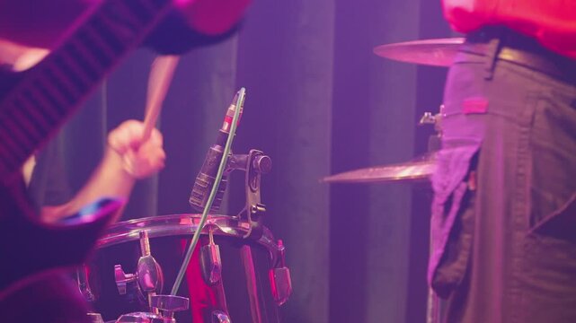 Drum kit with microphone in focus, blurred guitarist in background. Energetic atmosphere with red green stage lights. Concept of rock, metal, nightlife.Live music performance