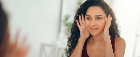 A young woman stands in front of a mirror while smiling. She touches her face with her hands and appears to be practicing different facial expressions. The setting is a bright room with simple decor. © Prostock-studio