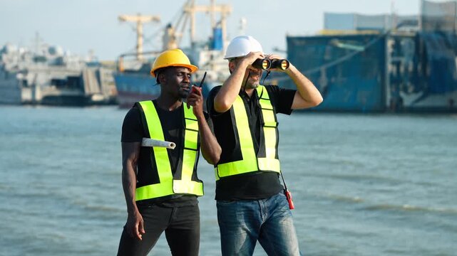 Two diverse port workers monitor shipyard activity. Engineer uses walkie talkie holding plans while colleague looks through binoculars. Industrial maritime logistics, safety gear, cargo vessels