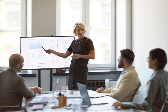 Businesswoman presenting sales data on monitor during team meeting, discussing strategy and performance metrics. Useful for corporate communication, analytics, leadership