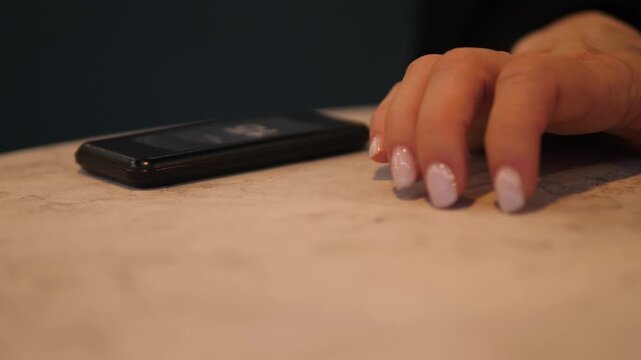 Close up of a woman's hand with manicure impatiently tapping fingers on a table near a smartphone. Female waiting for an important call or message in a cozy cafe.