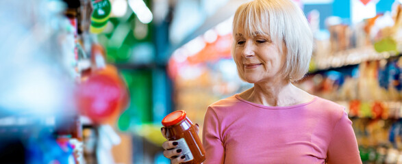 A woman is in a grocery store looking at a jar of sauce. She stands among shelves filled with products. Bright colors are around her, creating a lively shopping environment. © Prostock-studio