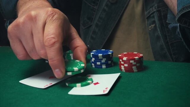 Male poker player places green and black chips on two playing cards during a game at a table with colorful poker chips in the background