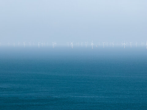 Aerial view of an offshore wind farm with rows of white wind turbines standing in the blue sea under a hazy sky in Plurien, Brittany, France.