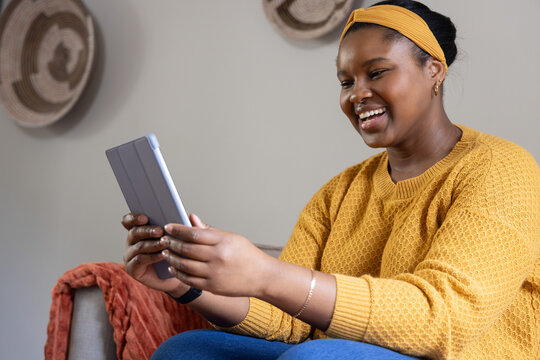 African American woman sitting at home holding gray tablet, wearing mustard sweater, orange throw