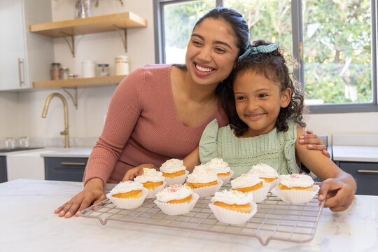 Mother and daughter in pink top, green dress smiling presenting cupcakes on cooling rack in kitchen