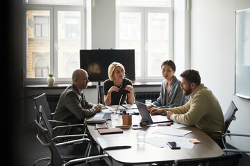 Obraz premium Wide shot of diverse business team meeting in office, discussing strategy around laptops and documents. Corporate planning concept for teamwork, leadership, consulting, communication