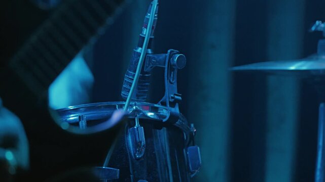 Close up detail of drum with microphone attached to rim. Scene is illuminated by dramatic blue stage lighting, creating intense atmosphere of heavy metal concert or rock show. Dark and moody