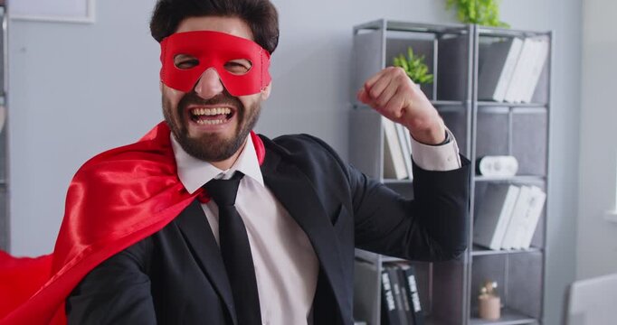 Happy excited young funny business man in superhero face mask and cloak sitting at the desk in office and making yes gesture rejoicing in successful job. 4k video. Slow motion video.