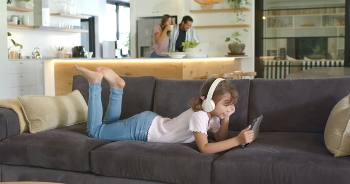 Family with teen girl lying on sofa wearing headset, using tablet to relax, parents prepping salad