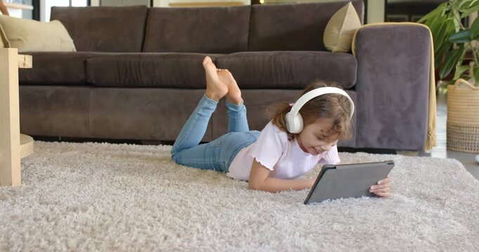 Female child wearing headphones lying on rug in living room, tapping tablet, laughing at funny clip