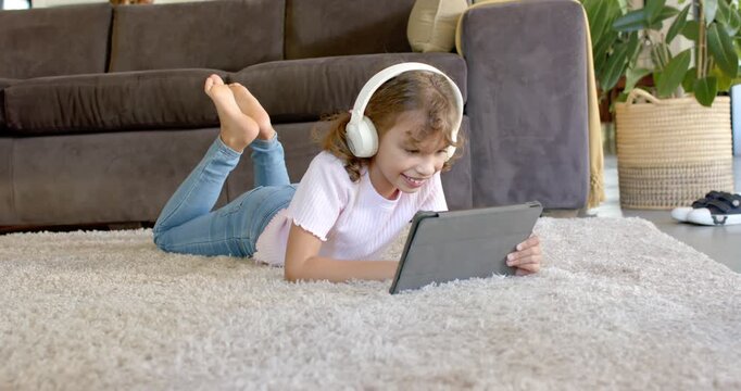 Female child lying on rug watching tablet for fun, content in headphones, tapping and smiling
