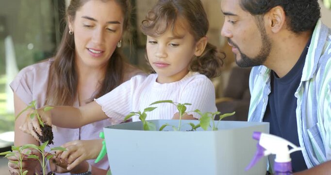 Diverse family teaching child as mom removing seedling from tray potting into gray planter on patio