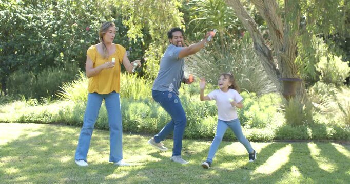 Diverse family playing mom lifting wand blowing bubbles, dad tossing bottle, child chasing in yard