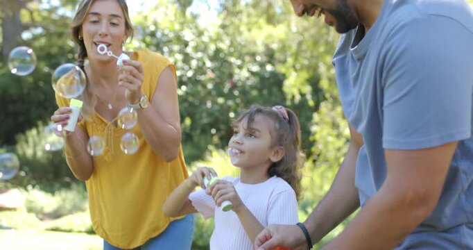 Family of 3 on sunlit lawn woman raising bottle dipping wand blowing bubbles teaching youth girl
