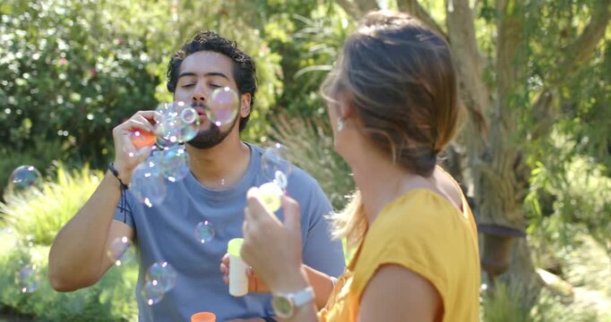 Couple taking turns blowing bubbles in sunlit garden, man raising wand initiating bottle reloading