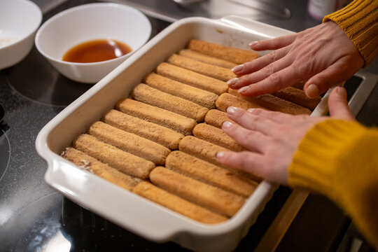 Close-up of hands layering ladyfingers for tiramisu in a ceramic baking dish on kitchen counter.